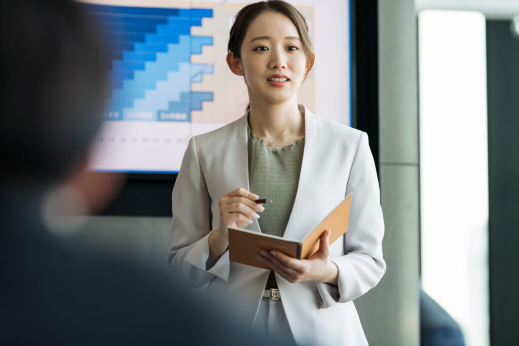 社内で同じ部署の方々に企画説明をしている白いスーツを着た女性の写真。キャリアコーチングmCalmee（カーミー）